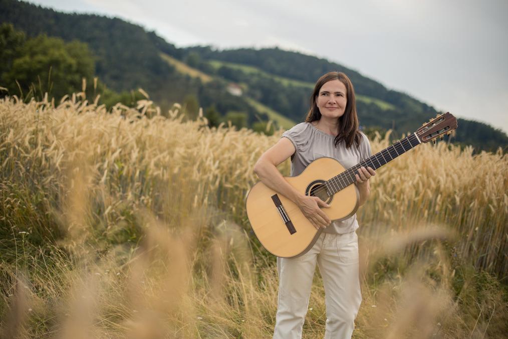 Eine Frau mit Gitarre steht vor einem Weizenfeld mit Bergen im Hintergrund.