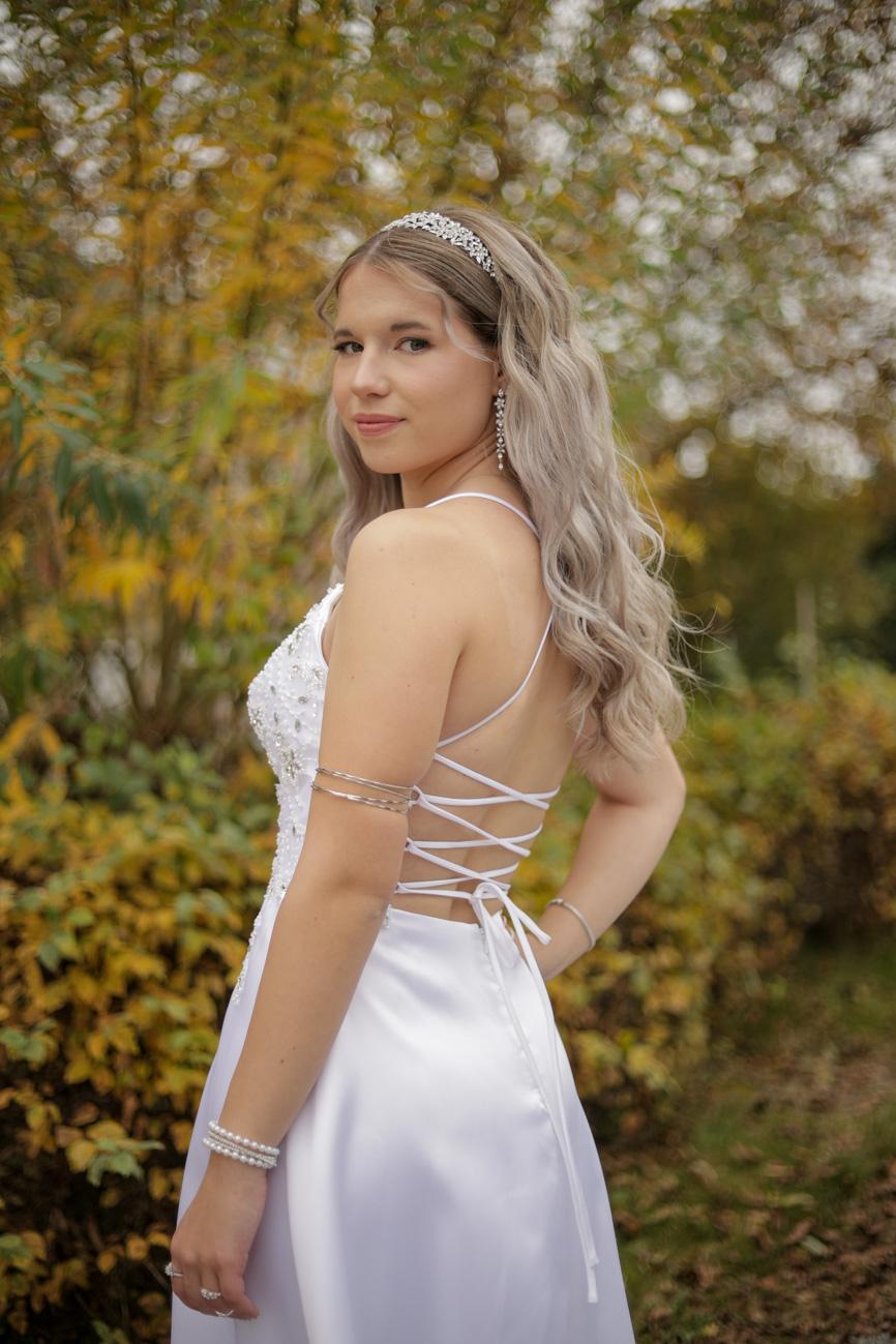 Eine Frau im Ballkleid schmunzelt in die Kamera mit Bäumen im Hintergrund.